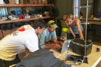 Three researches in field clothing, surrounded by equipment, one seated, gathered around a table in a science lab looking at a laptop.