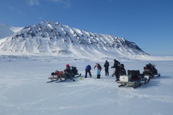 Team of scientists in winter clothing with snowbile in front of snow-covered arctic scene with mountain in background