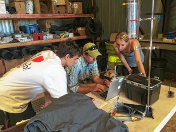 Three researches in field clothing, surrounded by equipment, one seated, gathered around a table in a science lab looking at a laptop.