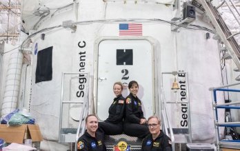 NASA crewmembers, in flight uniform, seated in front of NASA-white space capsule housed in aircraft hangar