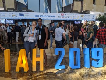 Candid photo of Sarah McKnight (left) and Marsha Allen (right) in front of crowd at a conference center, standing behind knee-high orange and blue block letters saying "IAH 2019"