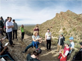 Dr. Sheila Seaman gesturing and teaching to group of students surrounding her on hillside of red-orange desert landscape with Saguaro cacti.