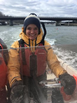 Portrait photo of Hannah Baranes piloting a small boat, smiling and dressed in yellow and orange foul weather gear.