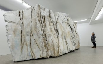large sculpture resembling calved ice block, in white room with gray-clad, long-haired person for scale. Image source: MassMOCA