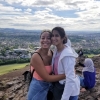 Raquel Bryant and Julie Beck facing camera in half-embrace atop rock outcrop in front of green pastoral scenery surrounding Edinburgh