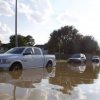 Pickup trucks and light SUVs parked to the side of a road flooded with brown water up to the bottom of the truck