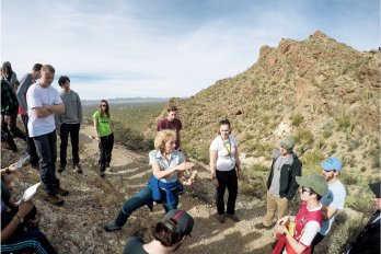Dr. Sheila Seaman gesturing and teaching to group of students surrounding her on hillside of red-orange desert landscape with Saguaro cacti.