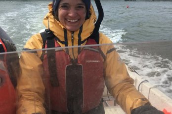 Portrait photo of Hannah Baranes piloting a small boat, smiling and dressed in yellow and orange foul weather gear.