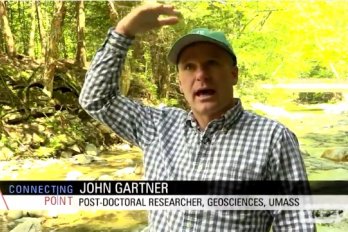 Screen capture of post-doc John Gartner speaking to camera in front of Chicklea River in Hawley, MA. John is gesturing to show height of floodwaters above his head.