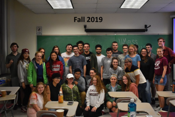 Over a dozen members of the U-Mass Geography club posing for a group photo within a classroom.