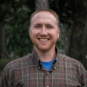 Portrait of Will Daniels, smiling, facing camera, in front of forested background