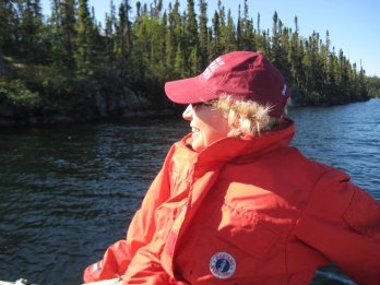Photo of the late Dr. Sheila Seaman, illuminated by sunlight and sitting reclined in a boat in a lake in the middle of a Canadian boreal forest, facing towards viewer but looking left, wearing sunglasses and field jacket / PFD. She is smiling.