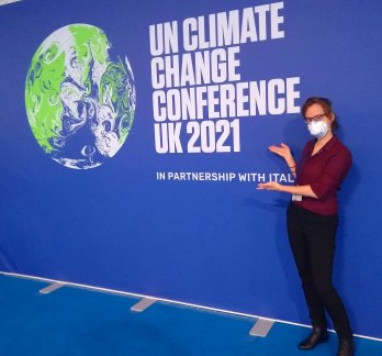 PhD Student Shaina Sadai stands in front of a blue wall with white letters and a globe on it.  The text reads: "UN Climate Chang