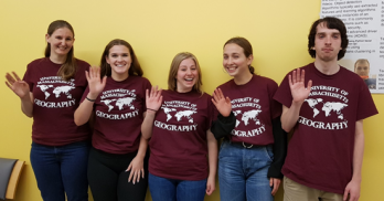 Five UMass Geography students standing in front of a yellow wall, wearing red t-shirts with U-Mass Geography logo, smiling waving at camera.