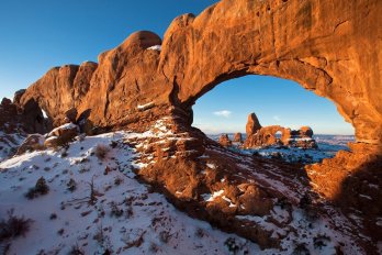 Orange sandstone rock arch in snowy desert landscape with clear blue sky (Turret Arch)