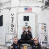 NASA crewmembers, in flight uniform, seated in front of NASA-white space capsule housed in aircraft hangar