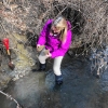 Dr. Julie-Brigam Grette, wearing bright-purple windbreaker and muck-boots, standing at muddy streambank, feet in water, examining hunks of clay-rich varve deposits she is holding. A shove rests, eager, on the bank.