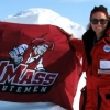 Photo of Dr. Julie Brigham Grette, smiling, wearing arctic cold-weather gear and sunglasses, holding U-Mass Minutemen flag in front of mountainous, snow-covered arctic landscape